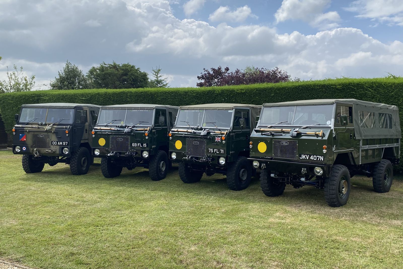 Four Land Rover 101 Forward Controls parked side by side on a lawn in front of a tall hedge