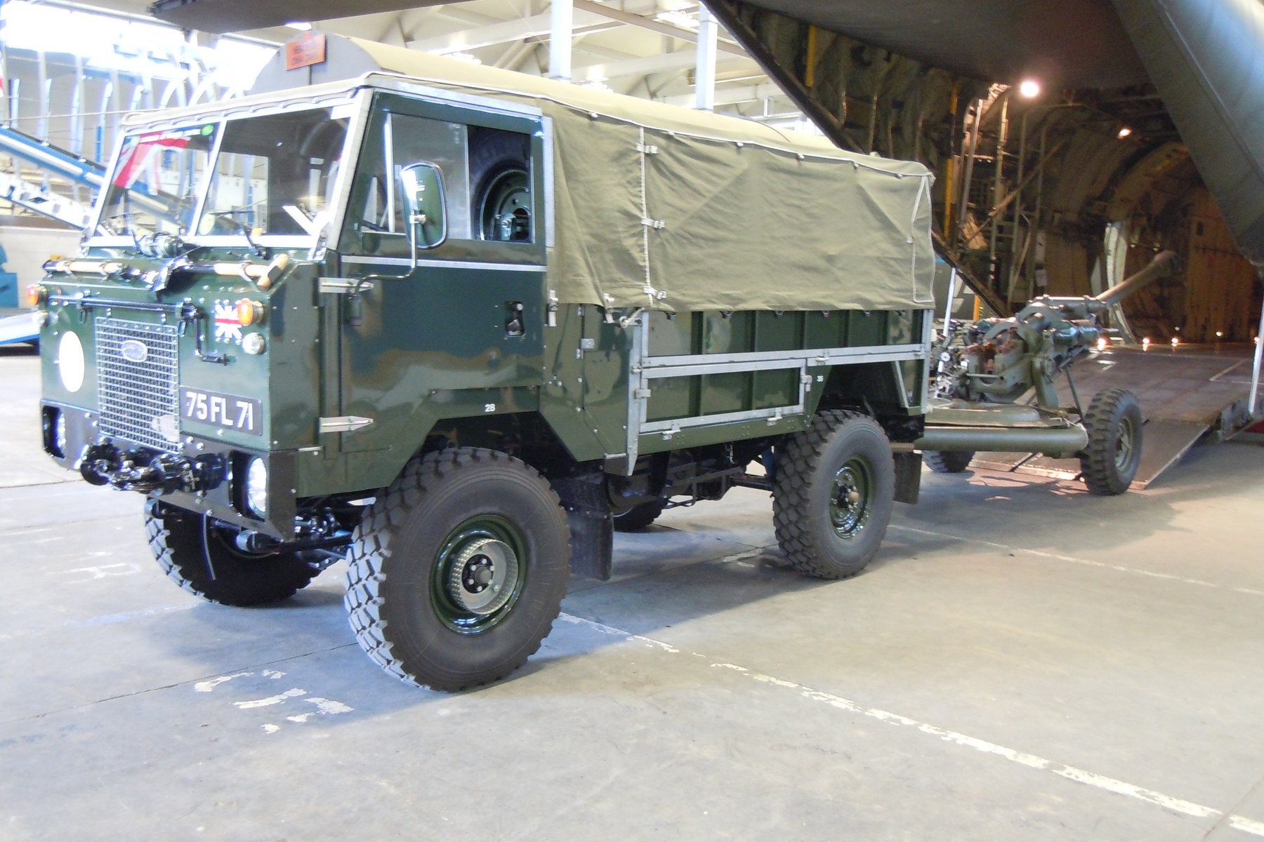 A green Land Rover 101 Forward Control with a field gun hitched behind it, beside the open rear ramp of a large aircraft